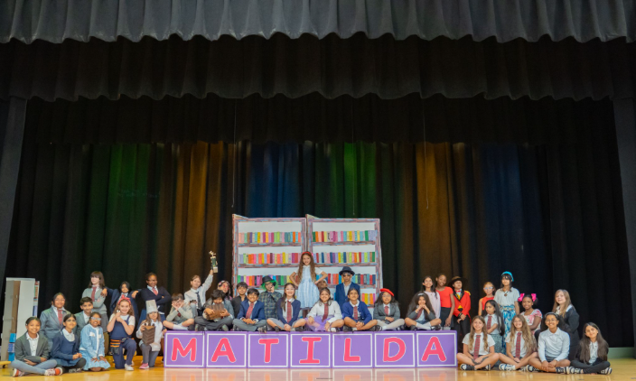 MON- A picture of the Matilda cast with the lead actors and actresses. It is on the stage with the black curtains. There are green, orange, and blue lights shining on the curtains. The background has painted books and boxes in purple with pink lettering of Matilda.