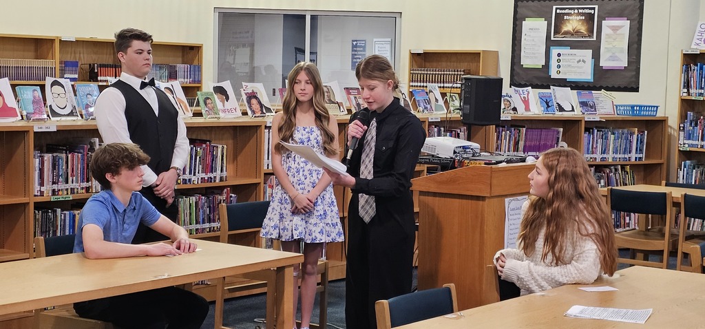 A girl speaking into a microphone and reading from a paper standing before a small group of students
