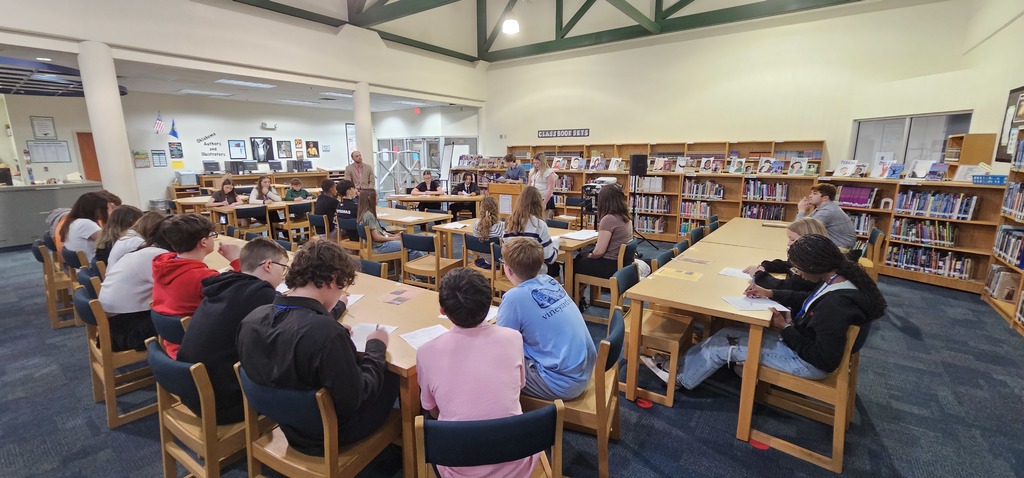 Students sitting at long tables in a library while a student presents from a podium