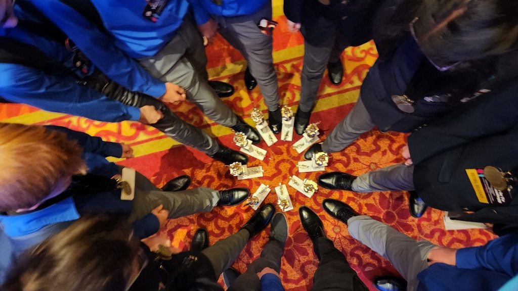Trophies are displayed in a circle on the floor. Students stand in a circle surrounding the trophies staring down at them.