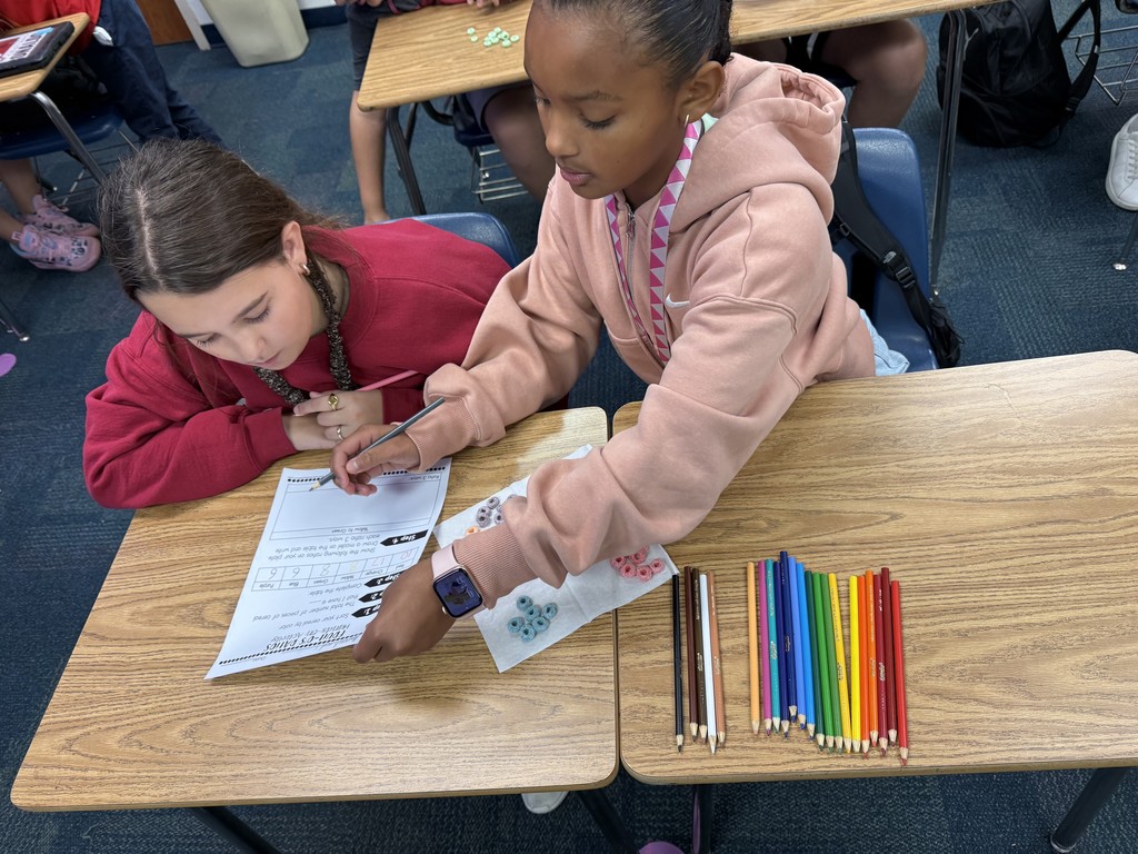 2 students looking at a worksheet with Fruit Loop cereal grouped by colors sitting on the desk beside them.