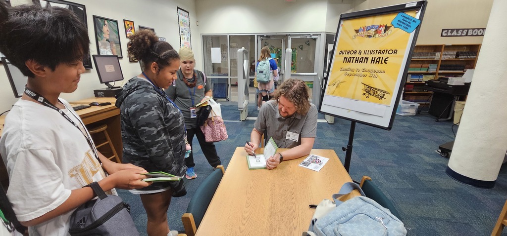 a boy and two girls standing in a line beside a table that author Nathan Hale is sitting at. They are talking with him as he autographs a book.