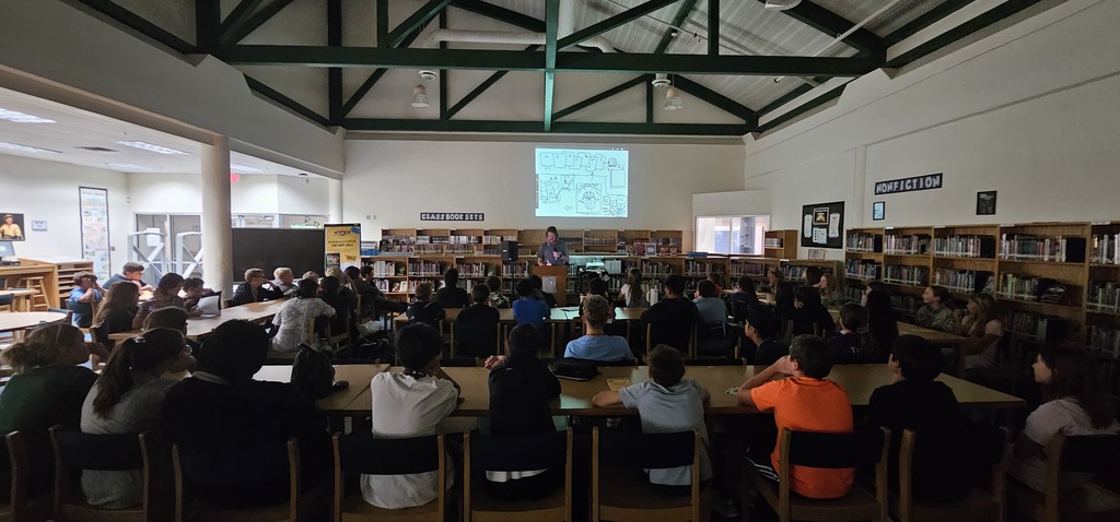 A group of students sitting at tables in a library watching author/illustrator Nathan Hale present while his drawings are being projected onto the wall