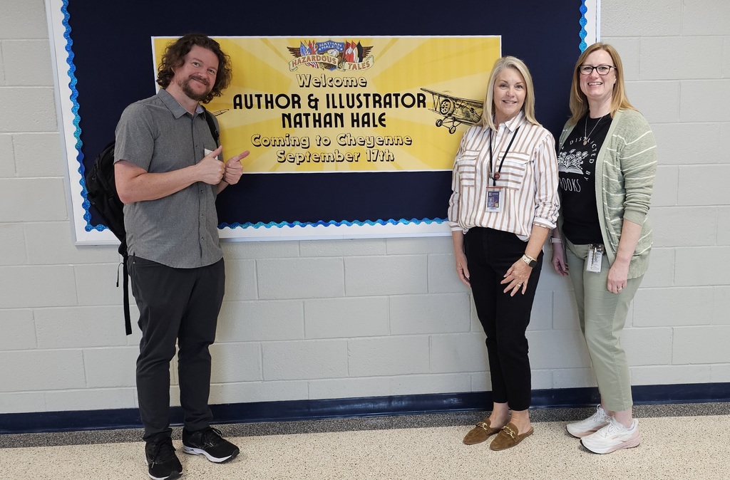 Author/illustrator, Nathan Hale, is standing in front of a bulletin board with a yellow poster on it. Two ladies are standing at the other end of the bulletin board smiling for the camera.