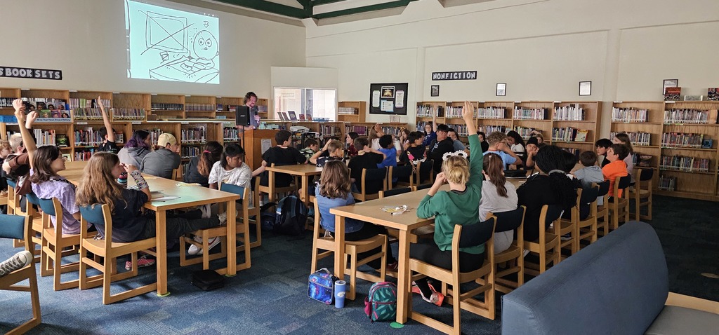 A group of students (many raising their hands), sitting at tables in a library watching author/illustrator Nathan Hale present while his drawings are being projected onto the wall