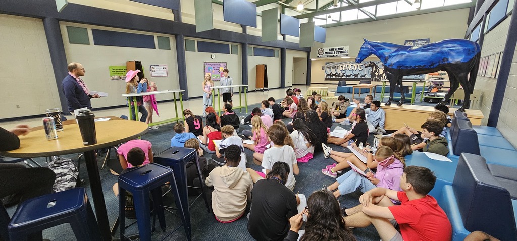 A group of around 50 students sitting on the floor in the commons while 4 students stand at the front of the group by two tall tables looking towards one another
