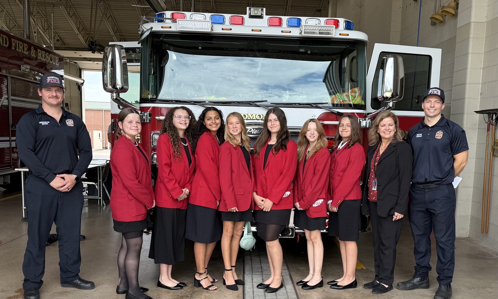 7 girls in red jackets and black skirts standing next to their teacher, Mrs. Wildman, and two firefighters in front of a fire truck. 