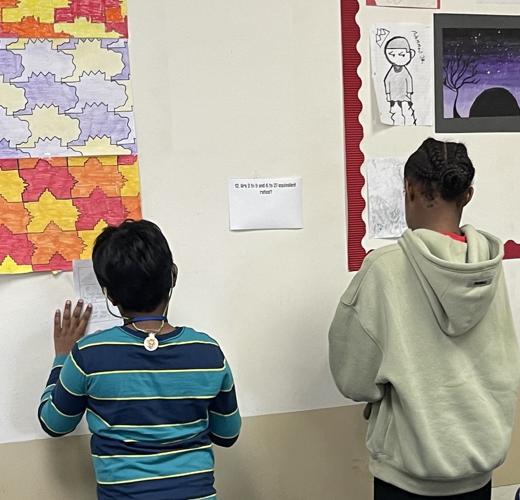 Two students work side-by-side on a math review activity working through a problem posted on the classroom wall.