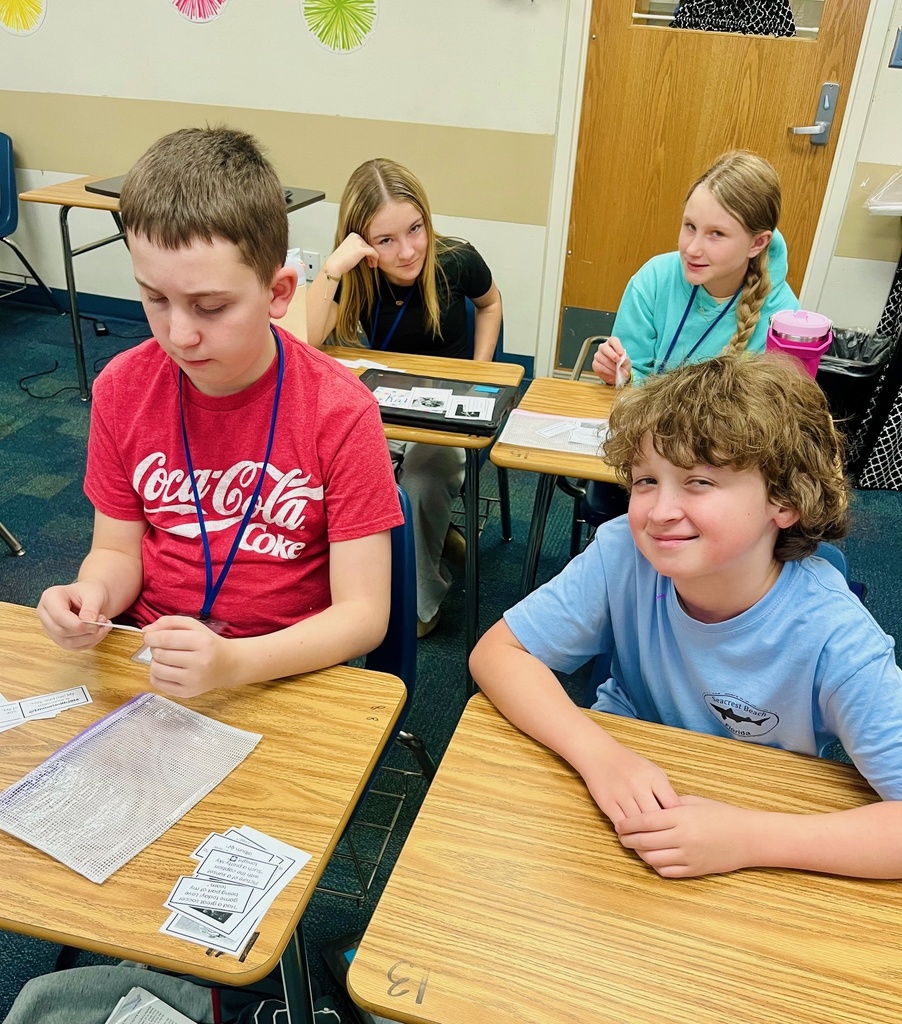 One student studies a social media example card while the student next to him smiles for the camera. Two students in the background also offer up smiles to the camera.