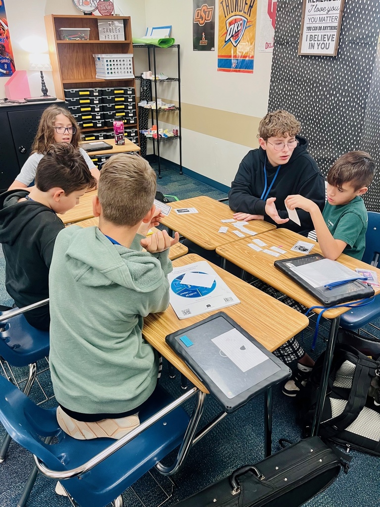 A group of 5 students are collaborating to determine which social media examples are safe to share while they sit together at a group of desks.