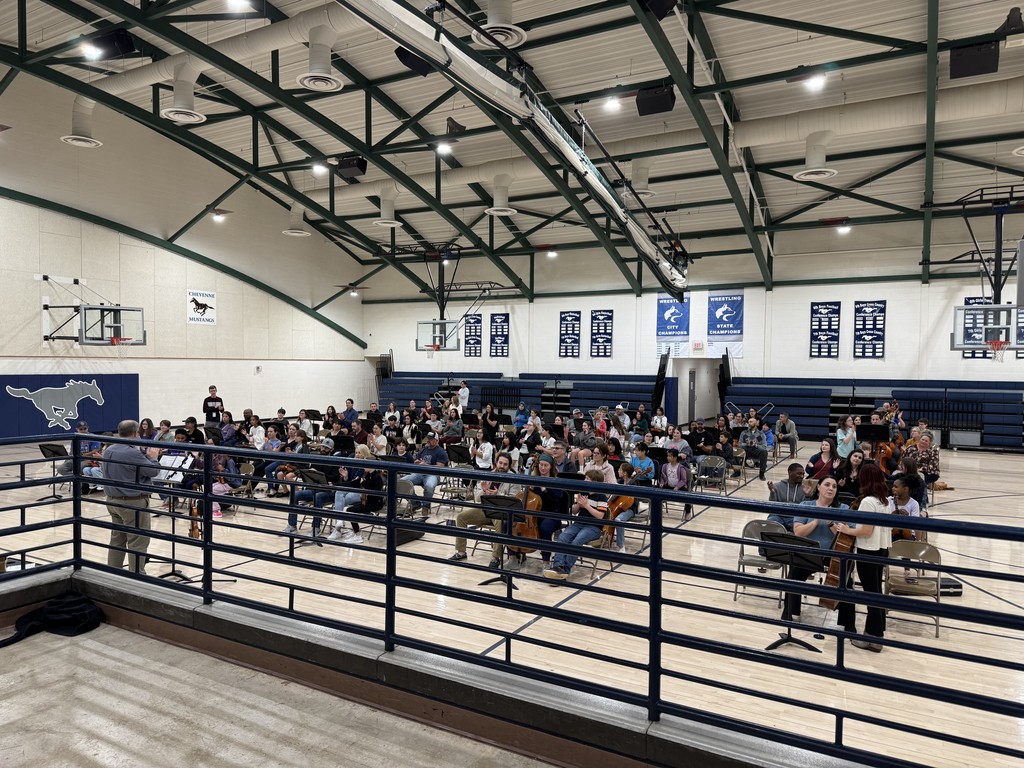 Mr. Ketch conducting with rows of people sitting in chairs with music stands in front of them. The student in the middle of each grouping is holding an orchestra instrument.