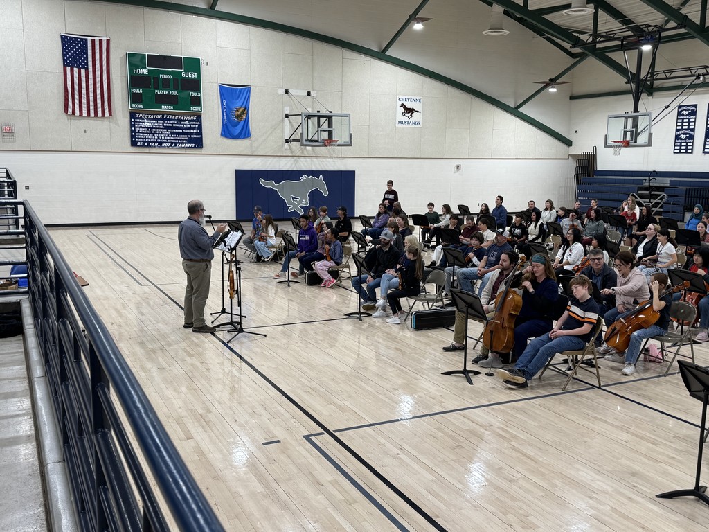 Mr. Ketch conducting with rows of people sitting in chairs with music stands in front of them. The student in the middle of each grouping is holding an orchestra instrument.