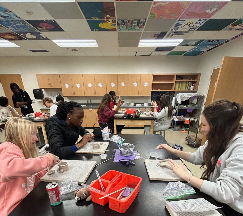 Students sit in a group at a table as they work on their clay pinch pot projects.