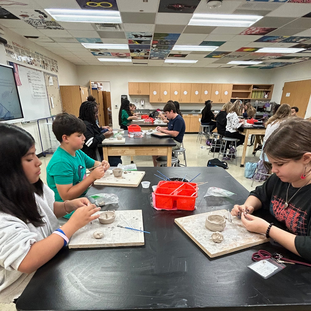 Students sit in a group at a table as they work on their clay pinch pot projects.