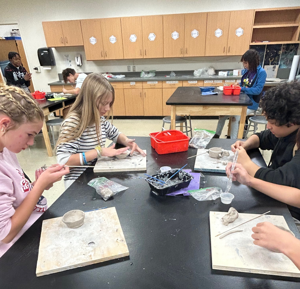 Students sit in a group at a table as they work on their clay pinch pot projects.