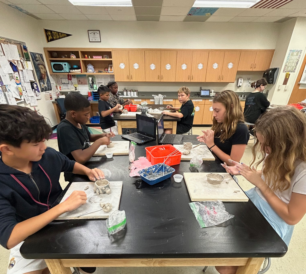 Students sit in a group at a table as they work on their clay pinch pot projects.