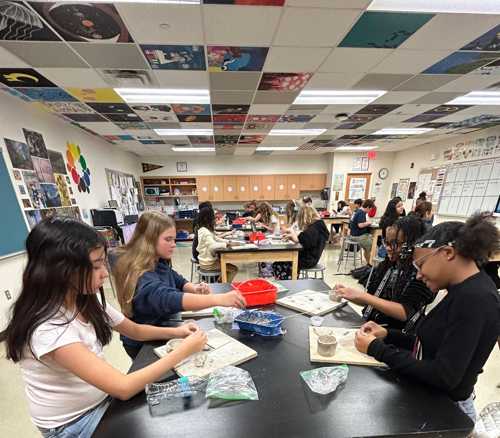 Students sit in a group at a table as they work on their clay pinch pot projects.