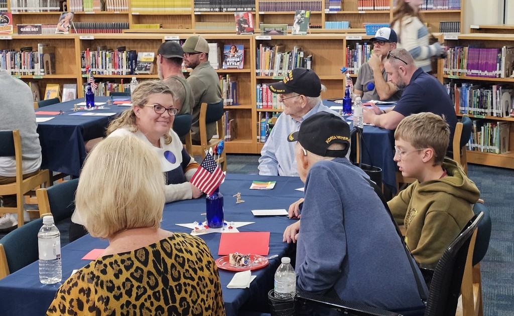 Two old men in wheelchairs wearing military hats sitting next to two ladies and a boy surrounded by others sitting in tables