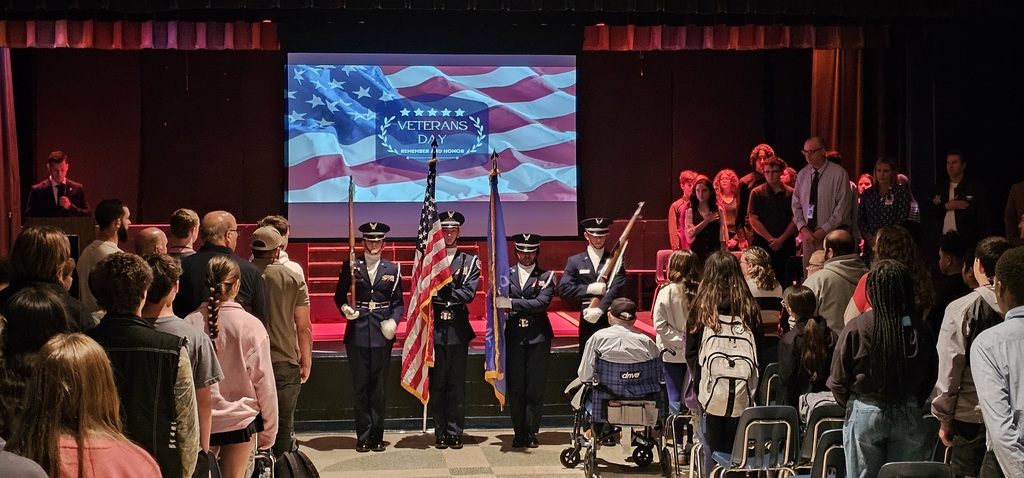 ROTC students holding presenting the US and Oklahoma flag while a group sits watching. There is a man on the stage talking into a microphone and a group on the other side of a projector screen standing.