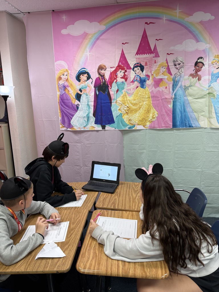 3 students wearing Mickey Mouse hats working on a worksheet while watching a chromebook. There is a Disney princess banner in the background.