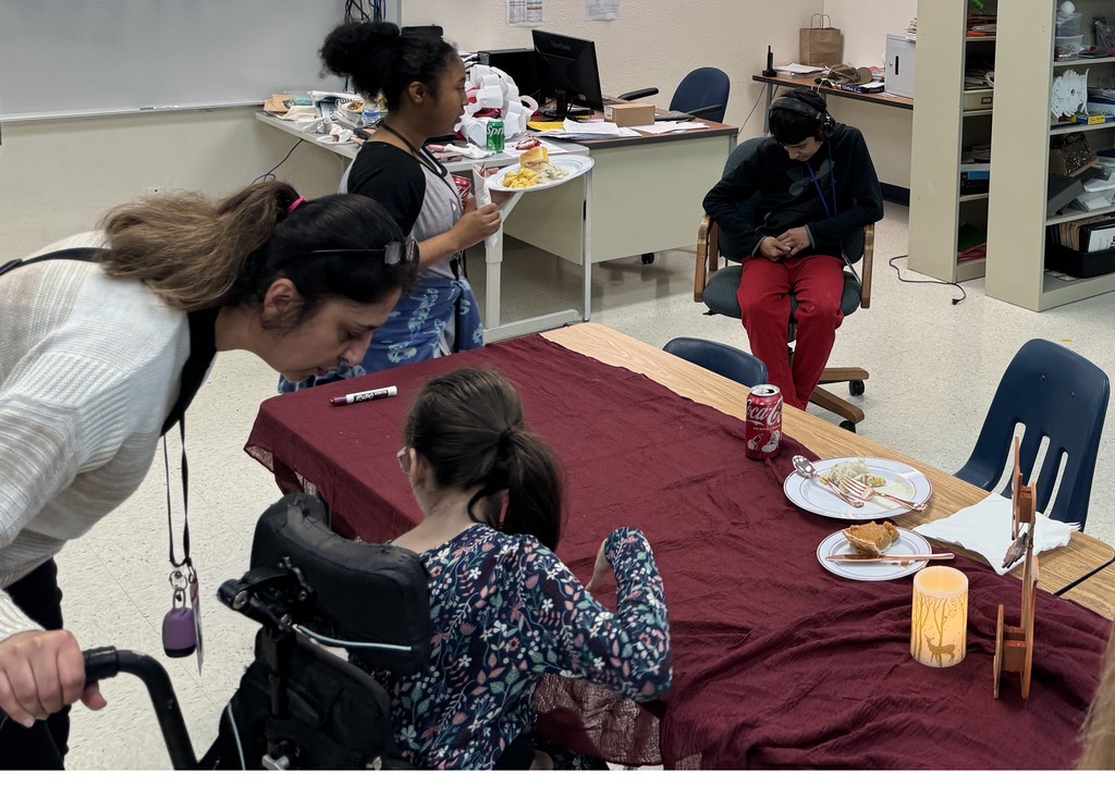 2 teen girls, 1 teen boy, and a woman near a table with a marron tablecloth, a slice of pumpkin pie, and a Coke.