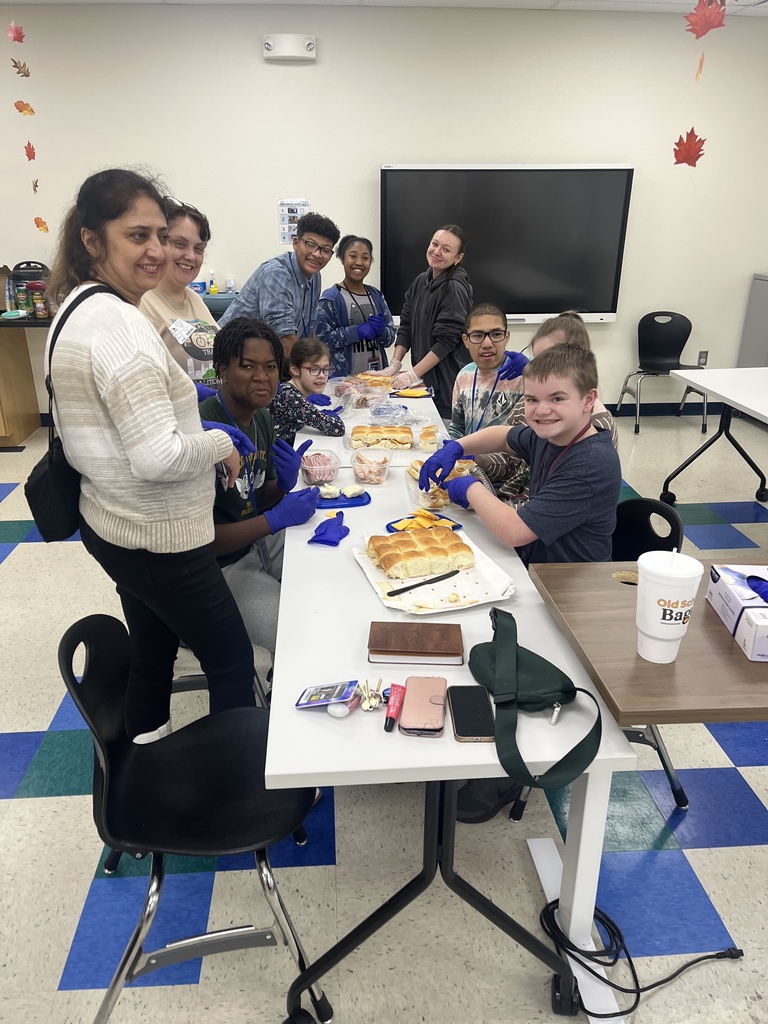 Students and classroom assistants sitting at two long tables making sandwiches