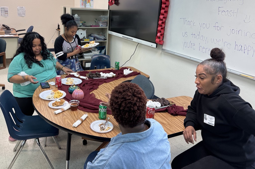 Three women and a teenage girl sitting at a table with Thanksgiving food on it, a marron tablecloth, and some ceramic pumpkins