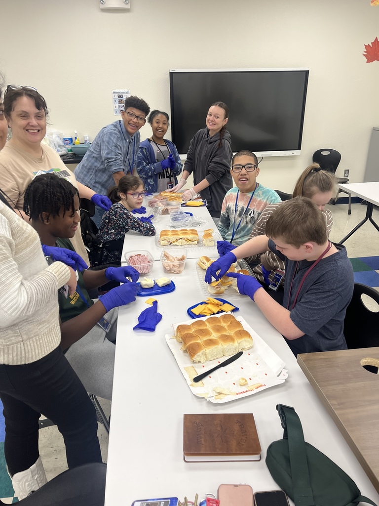 Students and classroom assistants sitting at two long tables making sandwiches
