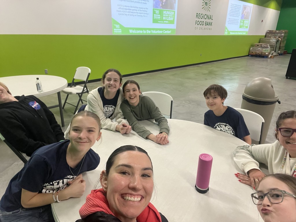 A group of students and 2 teachers taking a selfie while sitting around a white table with a pink water bottle in the middle