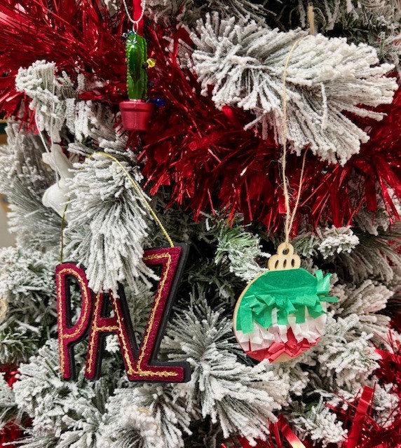 A close-up of a flocked Christmas tree with red garland, a small cactus ornament, a PAZ ornament, and a circular green, white, and red pianta style ornament