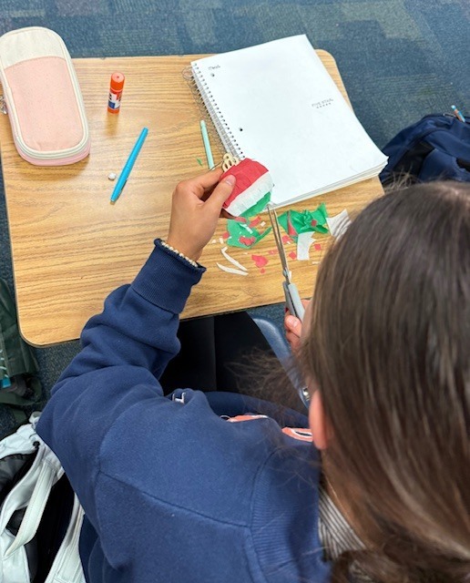 A student sitting at a desk with a pencil pouch, glue stick, notebook, and pen, cutting out a round red, green, and white ornament