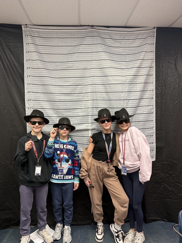 A group of 4 students wearing black detective hats and black sunglasses standing in front of a white and black line up height chart