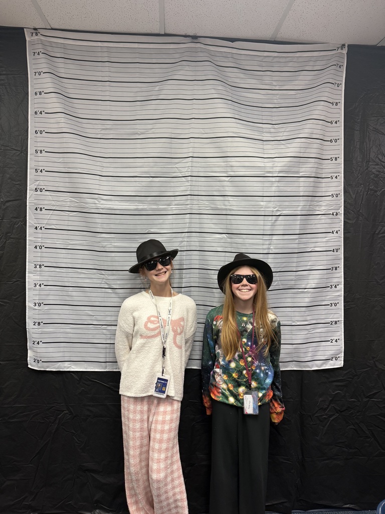 2 females wearing black detective hats and black sunglasses standing in front of a white and black line up height chart