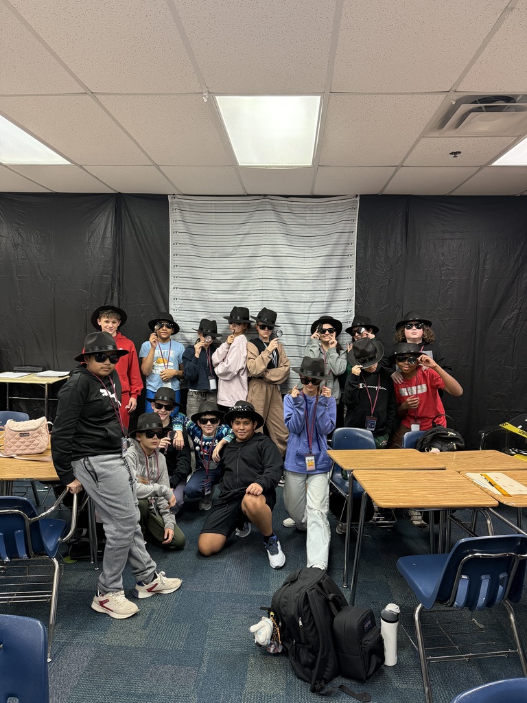 A group of 17 students wearing black detective hats, dark sunglasses, standing in front of a white and black height line up chart