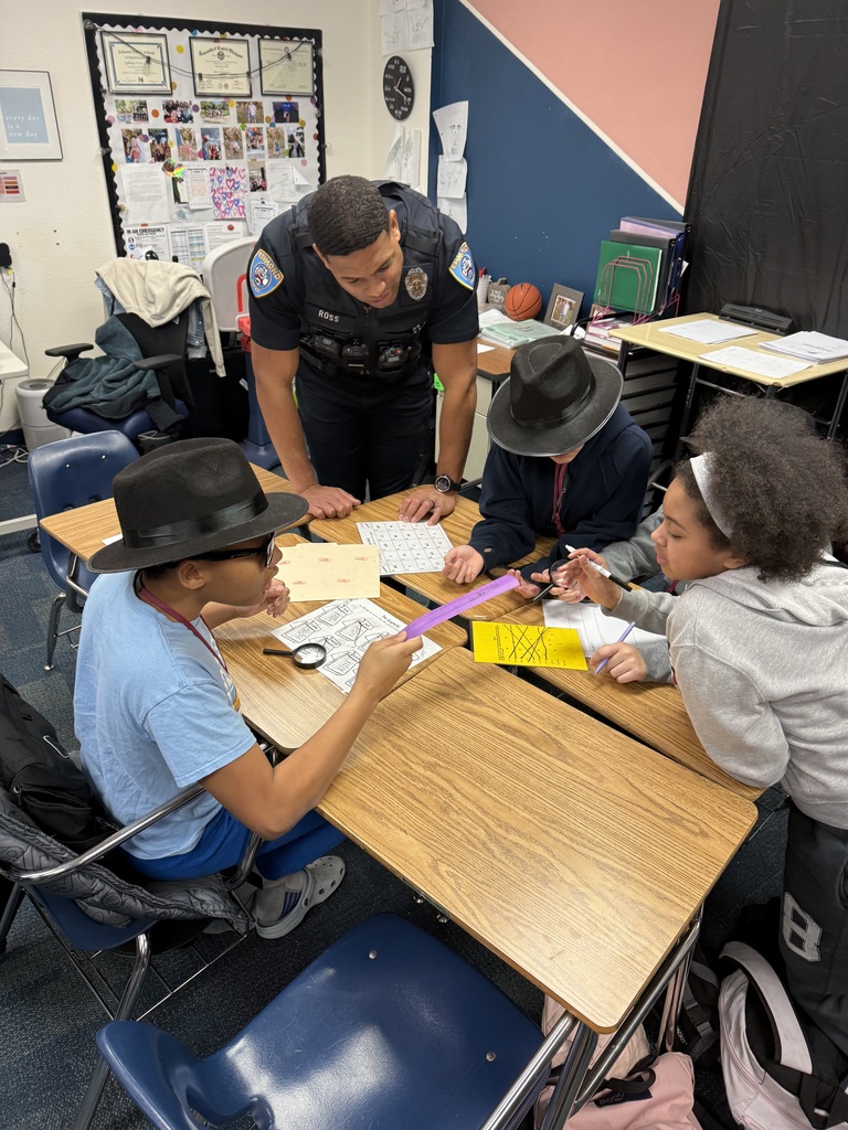 Officer Ross assisting 4 students who are sitting at desks. The kids are wearing old style detective hats and have a magnifying glass. 