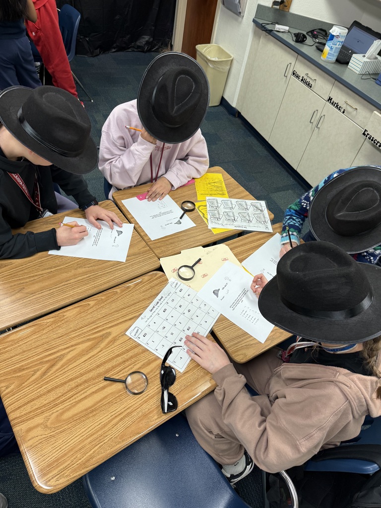 4 students filing out papers while sitting at desks. They are all wearing black detective hats and have magnifying glasses on the tables.