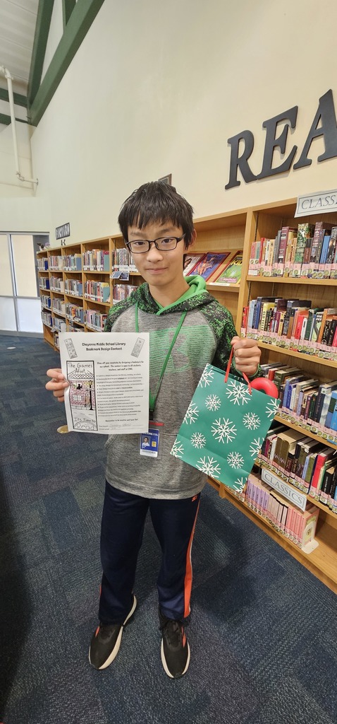 A student in a gray and green hoodie holding a green giftbag in one hand and his bookmark design form in the other hand