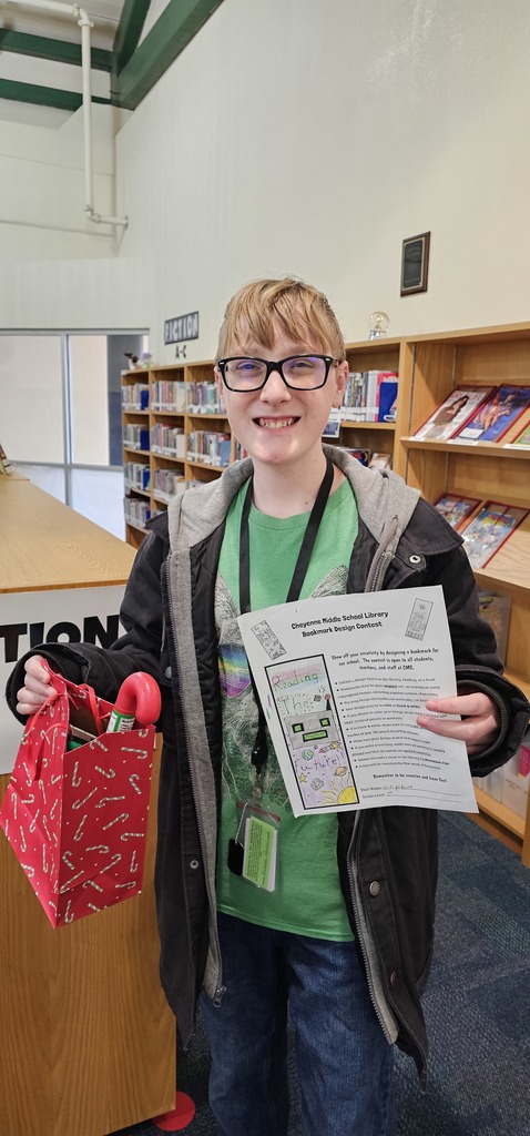 A student in a green shirt and black glasses holding a red giftbag in one hand and his bookmark design form in the other hand