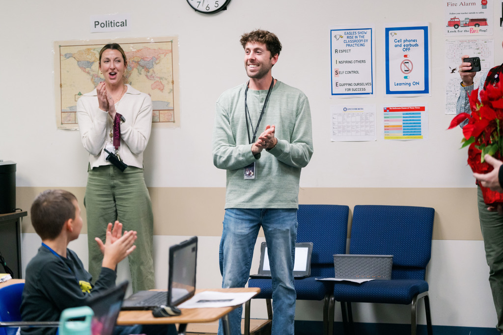 Daniel DeWindt, Summit Teacher of the Year, is pictured in his classroom smiling with hands clasped together as he learns that he is a finalist for District Teacher of the Year. A student and a Summit staff member can be seen smiling in the background of the picture.