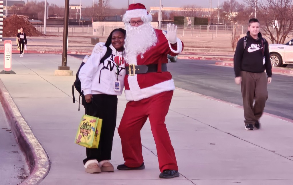 A person dressed as Santa posing with a girl carrying a Sour Patch tote with two students in the background walking to school with backpacks on their backs