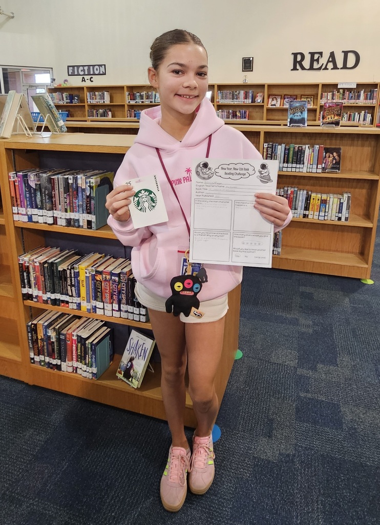 A girl in pink clothing holding a Starbucks gift card and a worksheet standing in front of library shelves.