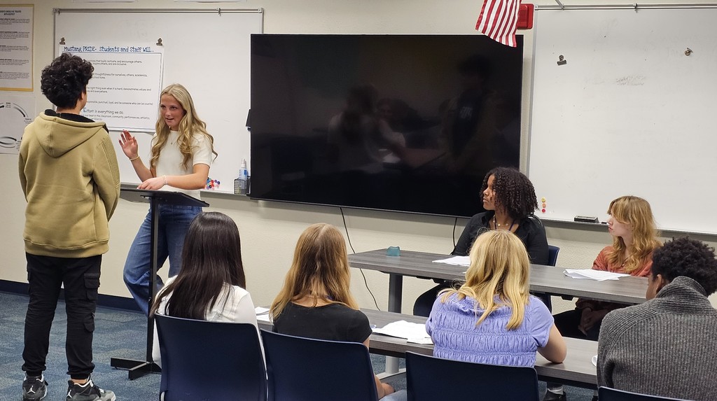 Students sitting in a mock courtroom with a girl holding up her right hand with a student bailiff standing in front of her
