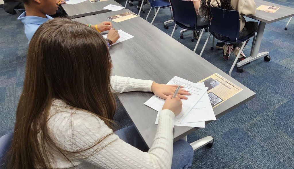 Students sitting at tables writing on a worksheet with a flyer that looks like an old time newspaper sitting on the table