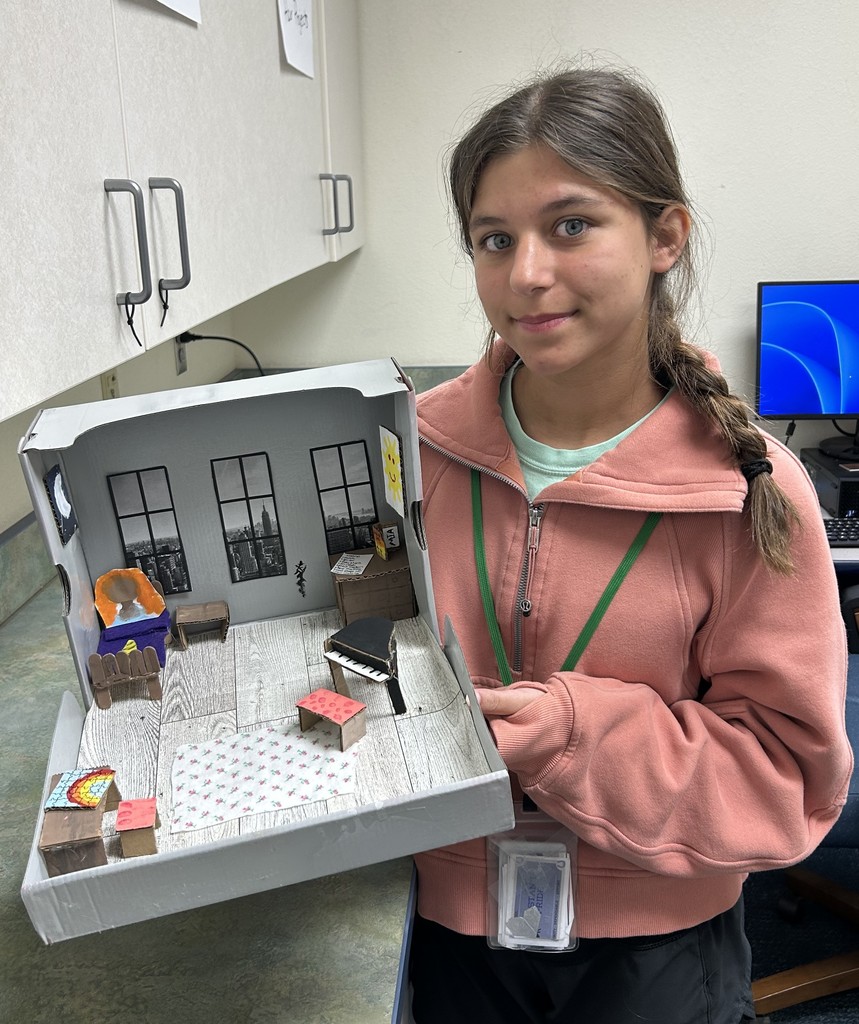 A teen girl holding a diorama of a room with a piano and windows overlooking NYC