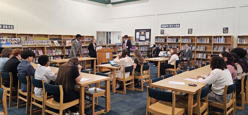Students dressed up sitting in the library that has been rearranged to look like a courtroom. 1 student is talking into a microphone while the judge looks on