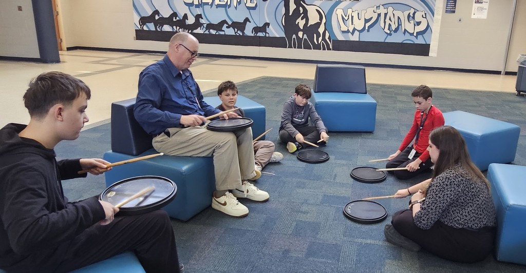 4 boys, Mr. Wheeler, and Mrs. Epperly-Reiswig holding drum sticks and black drum heads sitting in the Commons on the floor and/or blue seats