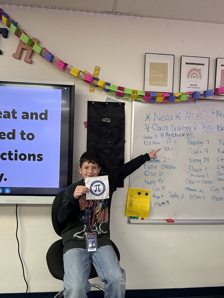 A boy holding a piece of paper with a Pi symbol on it pointing to a white board