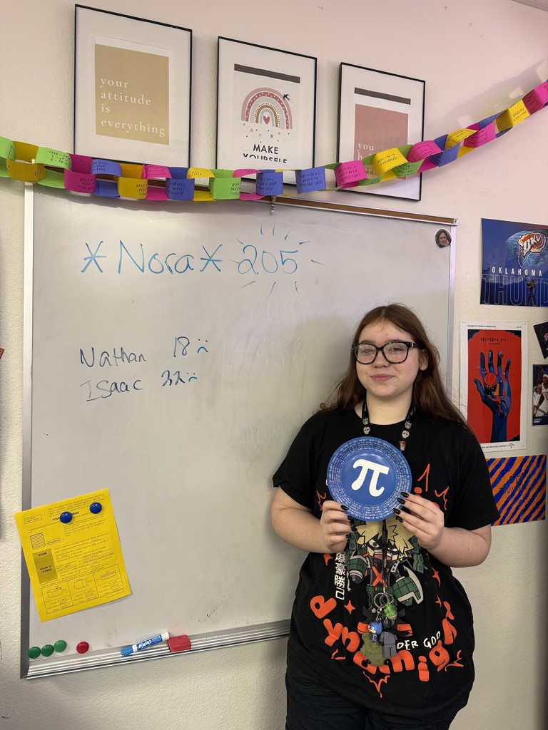 A girl standing by a white board with the words, "Nora 205." She is holding a plate with the Pi symbol on it.