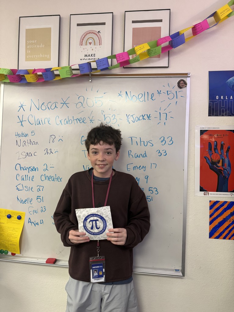 A boy standing in front of a white board holding a piece of paper with the Pi symbol on it.