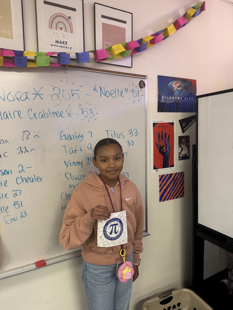 A girl in front of a white board holding a piece of paper with the Pi symbol on it.